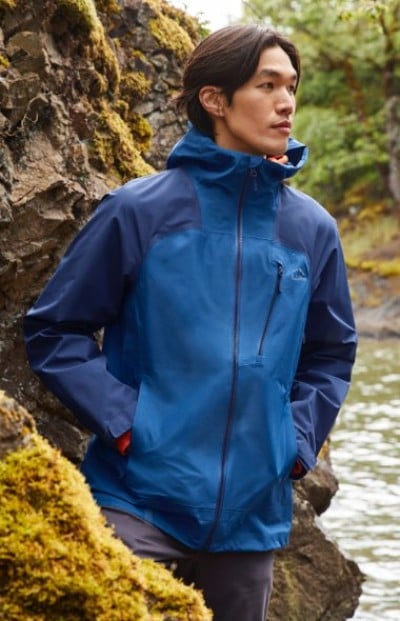 Person stands by mossy rocks and water, wearing a blue colorblock rain jacket with hands in pockets.
