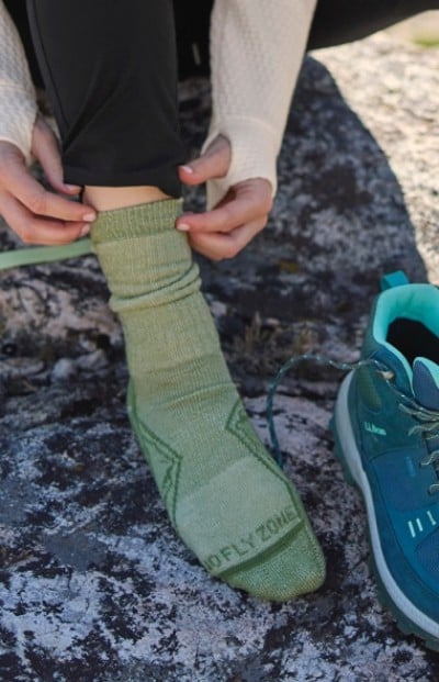 Woman sits on a rock adjusting a green hiking sock on her left foot.