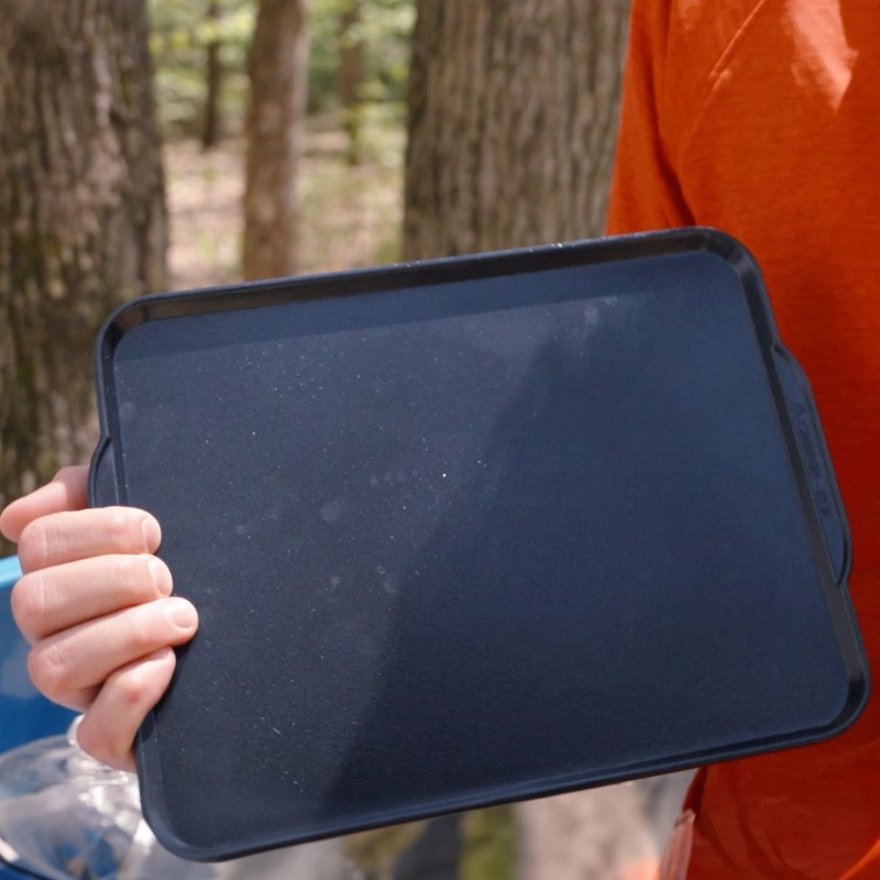 A person in an orange shirt holding a black griddle outdoors, possibly for cooking or grilling.