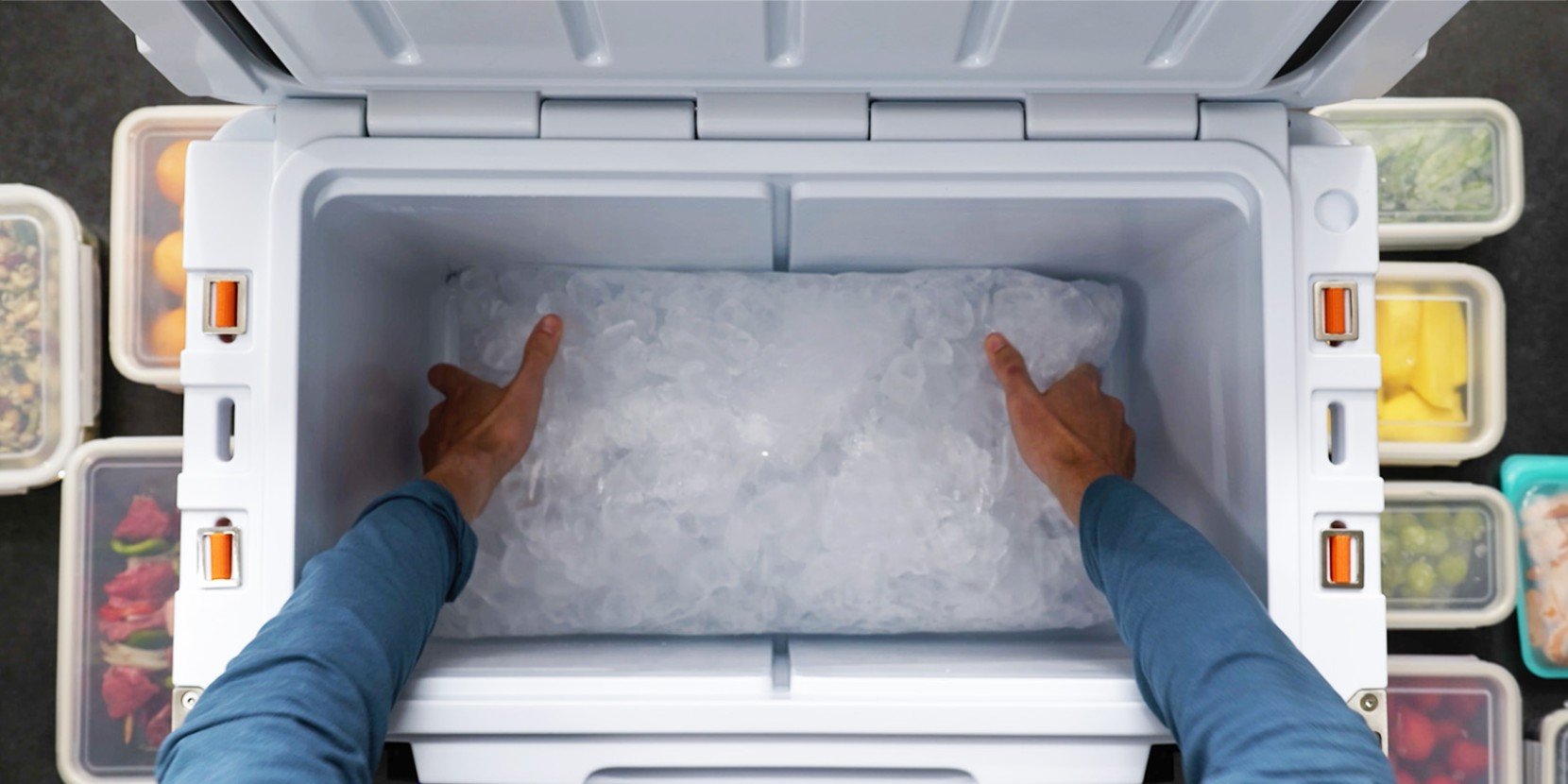 Hands are packing ice into a cooler, surrounded by various food containers, viewed from above.