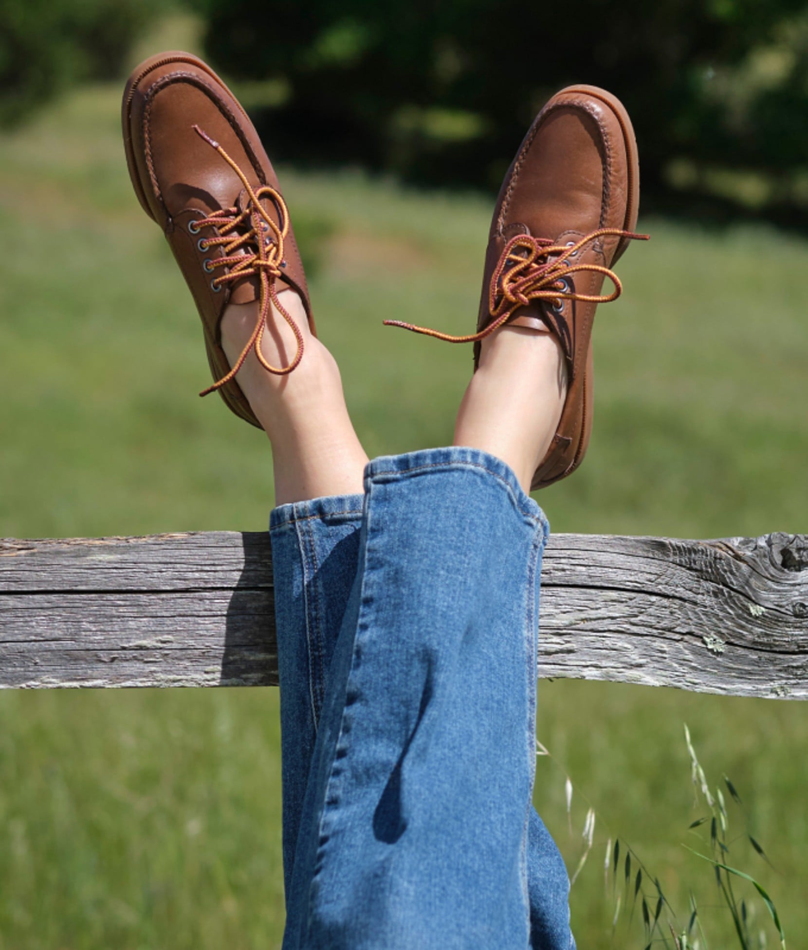 Person in brown leather moc-toe shoes propped on a wooden fence rail.