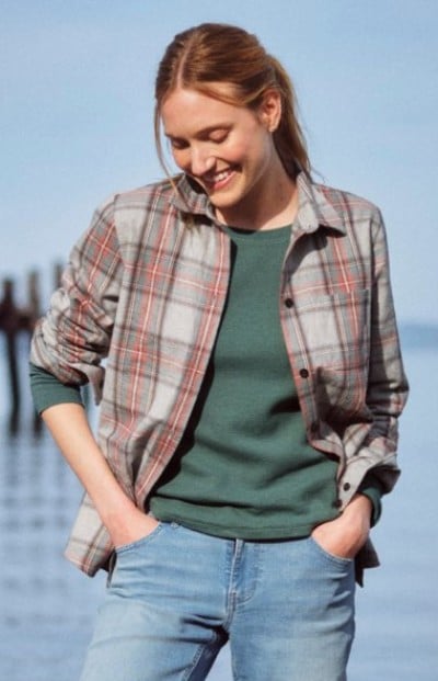 Person wearing a plaid flannel shirt over a green top standing outdoors with hands in pockets.