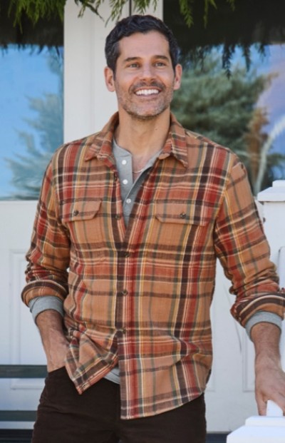 Man in plaid shirt stands outdoors, smiling with one hand in pocket and other on white railing, with trees in background.