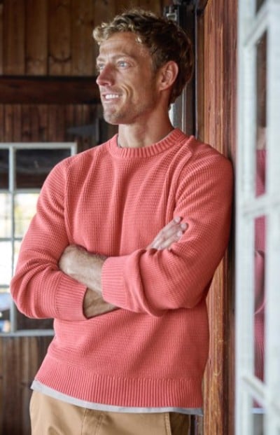 Person wearing a textured coral sweater and tan pants, standing indoors with arms crossed against a wooden wall.
