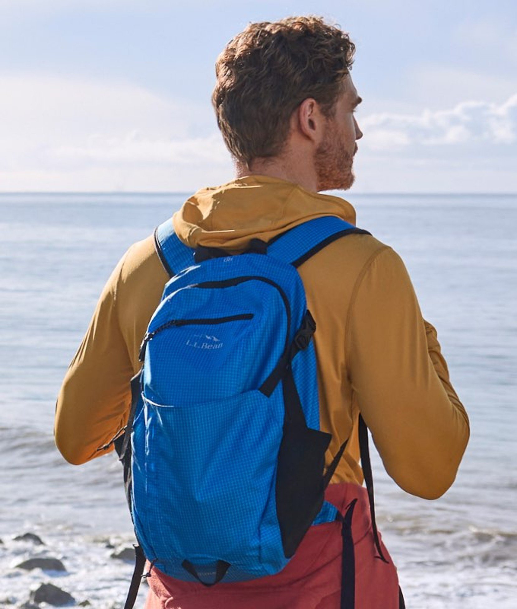 Person wearing a backpack and jacket, standing on a beach and looking toward the ocean. 