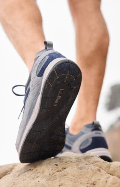 Athletic shoes stepping onto a rock, showing the tread and part of the legs against an outdoor backdrop.