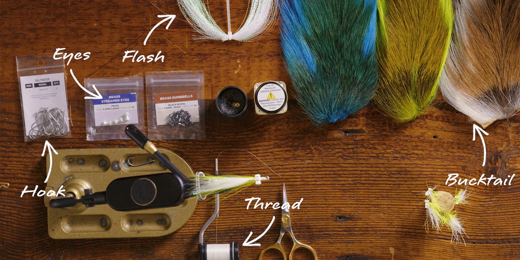Fly tying materials including hooks, eyes, flash, thread, bucktail, and a vise on a wooden table.