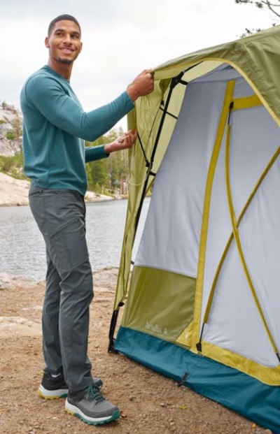 Person setting up a green and yellow tent by a lakeside, wearing a teal long-sleeve shirt, gray pants, and hiking shoes.