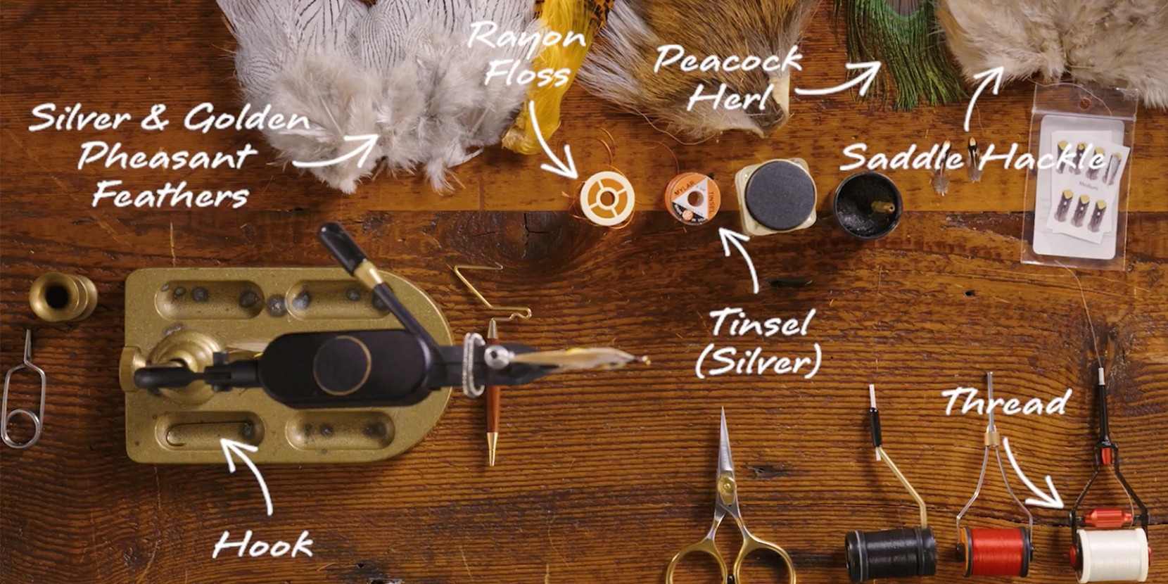 Fly tying setup with silver and golden pheasant feathers, rayon floss, hook, and tools on a wooden surface.